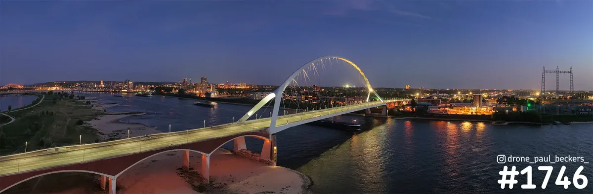 Panorama van De Oversteek over de Waal in Nijmegen in de avondschemering, met verlichte boogbrug en de stad op de achtergrond