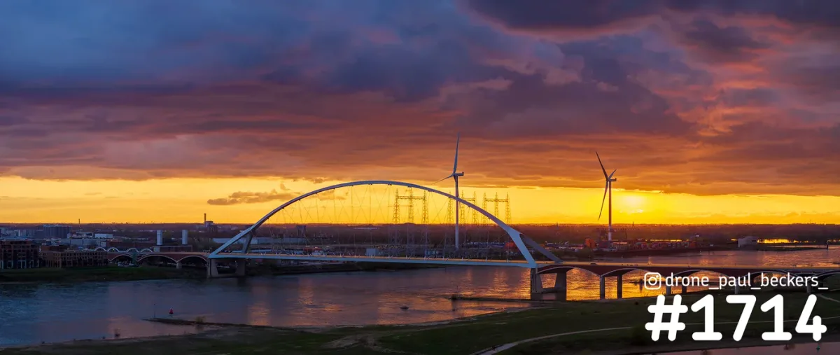 Luchtfoto van De Oversteek over de Waal bij Nijmegen tijdens zonsondergang, met windmolens en hoogspanningsmasten op het industrieterrein