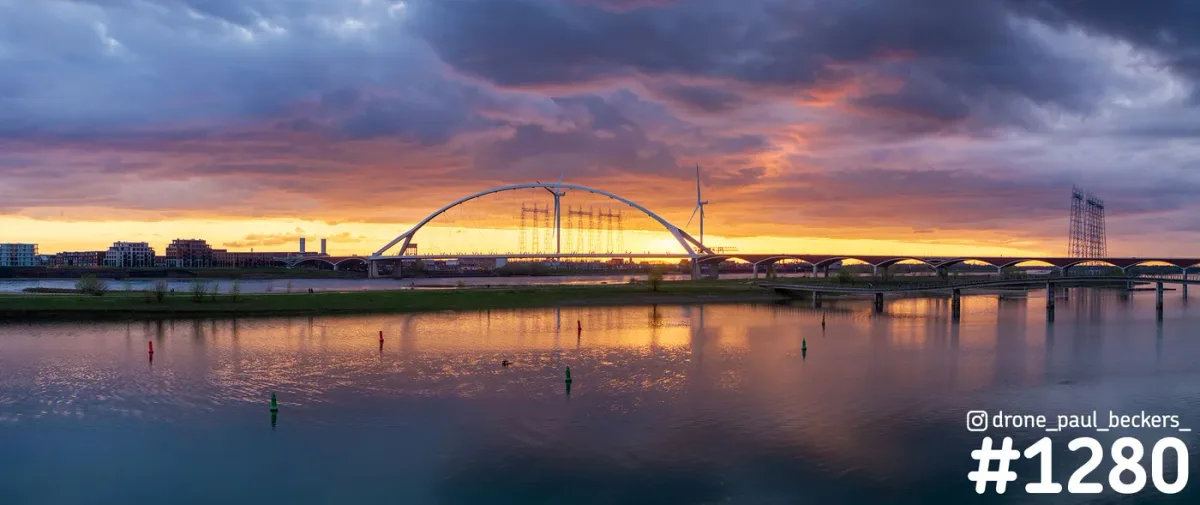 Panorama van De Oversteek in Nijmegen bij zonsondergang, met de boogbrug en oranje wolken weerspiegeld in de Waal