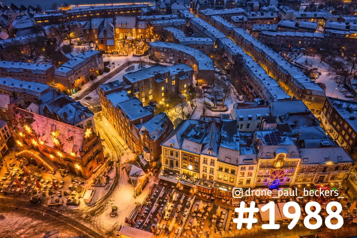 Luchtfoto van de Grote Markt in Nijmegen in de sneeuw, met verlichte gevels en terrastafels in het avondlicht