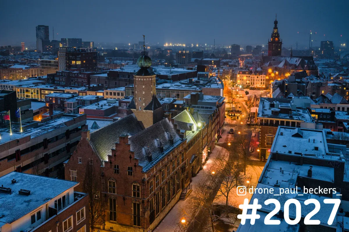 Luchtfoto van besneeuwd centrum Nijmegen in de avond, met trapgevel bij de Grote Markt en de Stevenskerk op de achtergrond