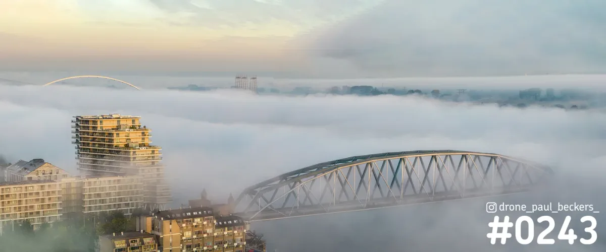 Luchtfoto van De Handelskade in Nijmegen: stalen boogbrug en appartemententoren in de ochtendmist, witte boog aan de horizon