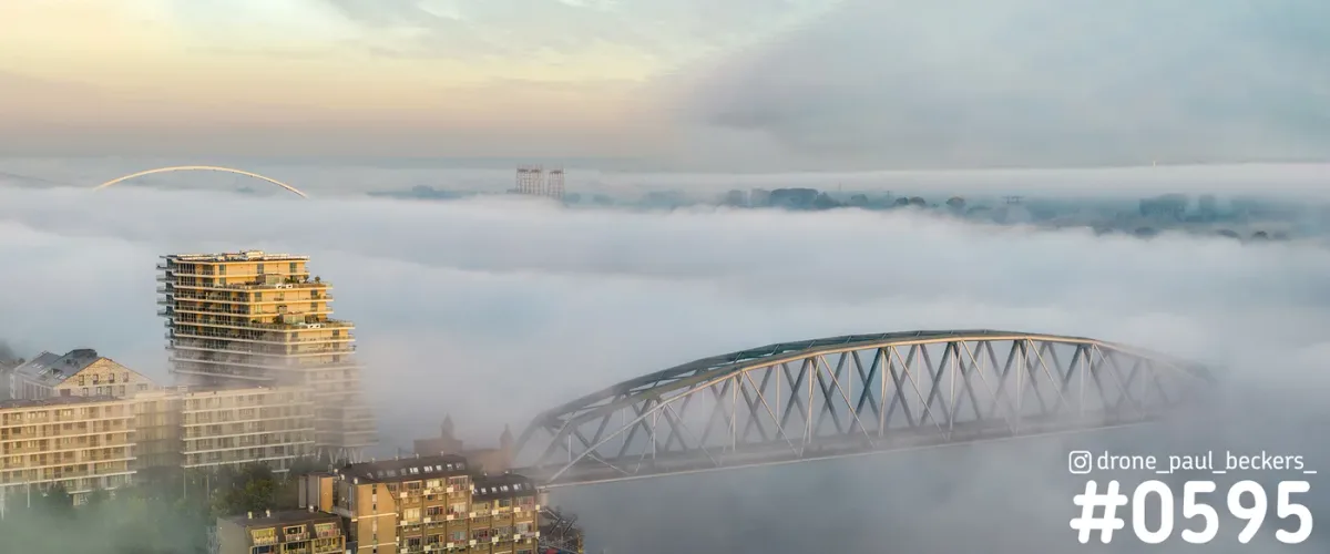 Luchtfoto van De Handelskade in Nijmegen: hoogbouw en een stalen boogbrug steken uit een dik mistdek in het ochtendlicht