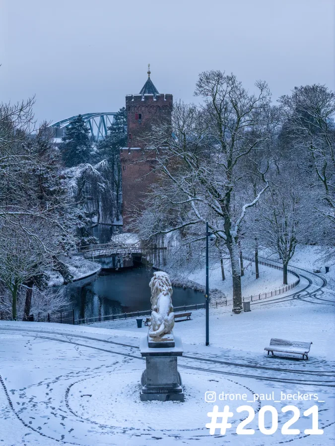 Kronenburgerpark in Nijmegen onder sneeuw, met stenen leeuw, rode baksteentoren en besneeuwde bomen langs de vijver