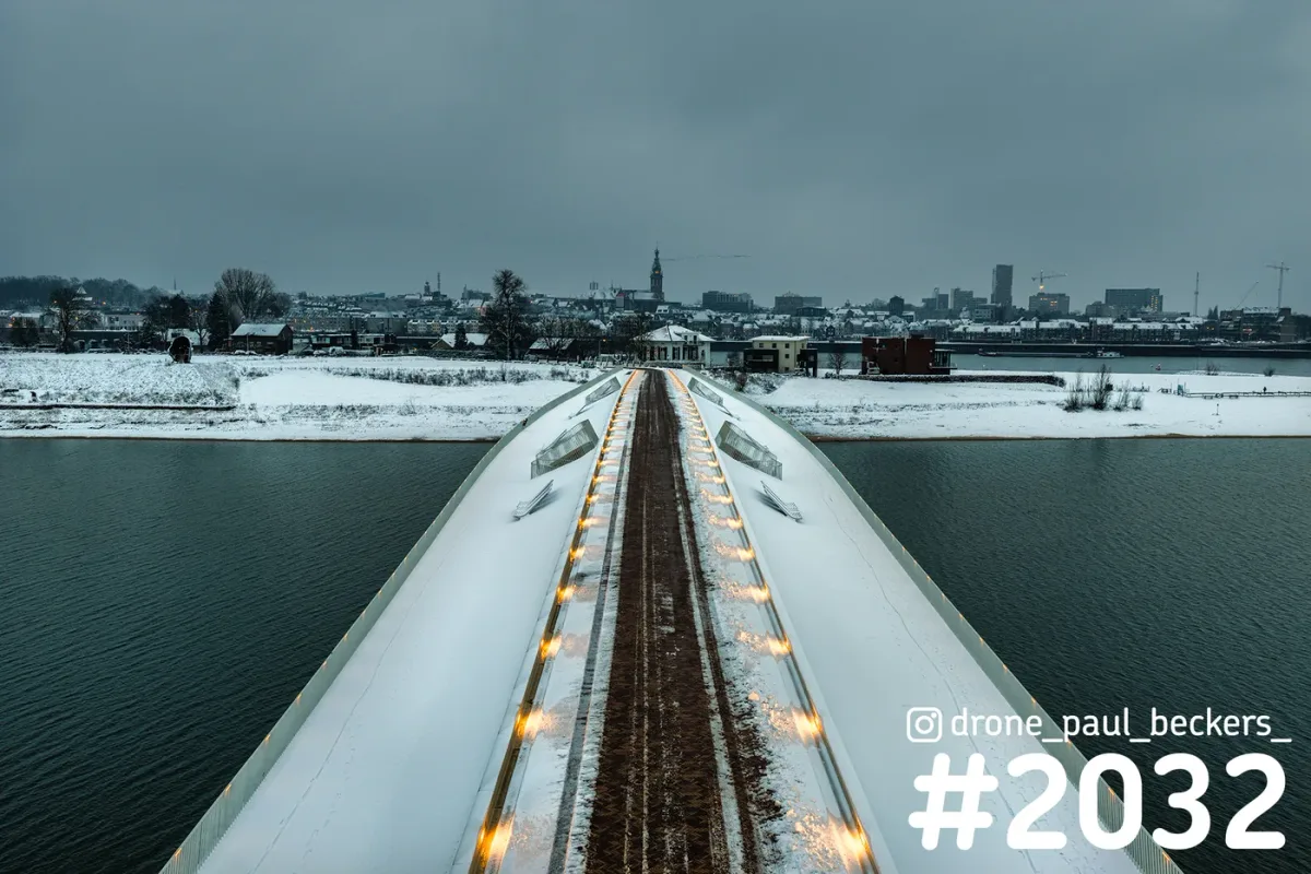 Luchtfoto van een besneeuwde brug over de nevengeul bij Lent, met de Stevenskerk van Nijmegen in de grijze winterlucht