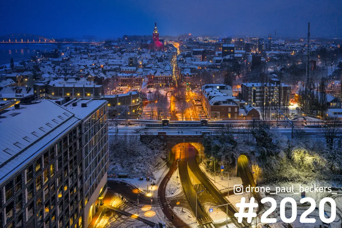 Luchtfoto van Nijmegen in de sneeuw tijdens het blauwe uur, met verlicht spoorviaduct en de Stevenskerk in de verte