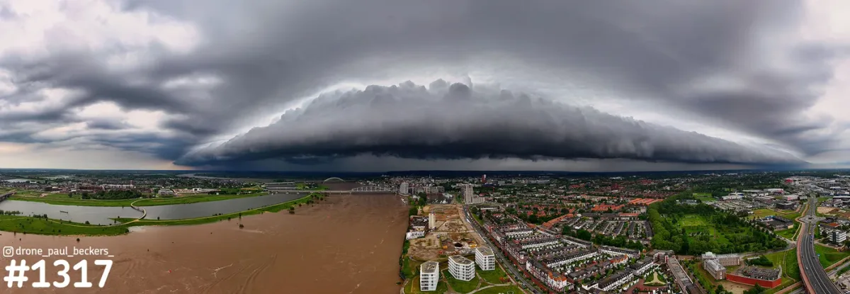 Panorama-luchtfoto van een brede onweerswolk boven Nijmegen, met de bruine Waal en de Nevengeul bij Lent links