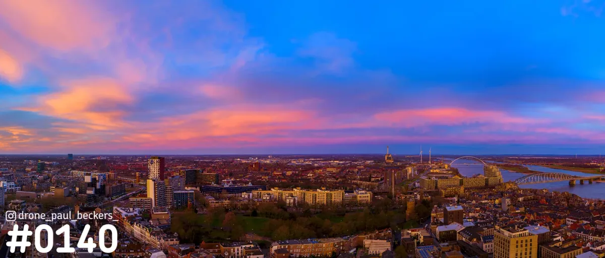 Luchtfoto panorama van Nijmegen in de avondschemering met roze en oranje wolken, de Waal en De Oversteek aan de horizon