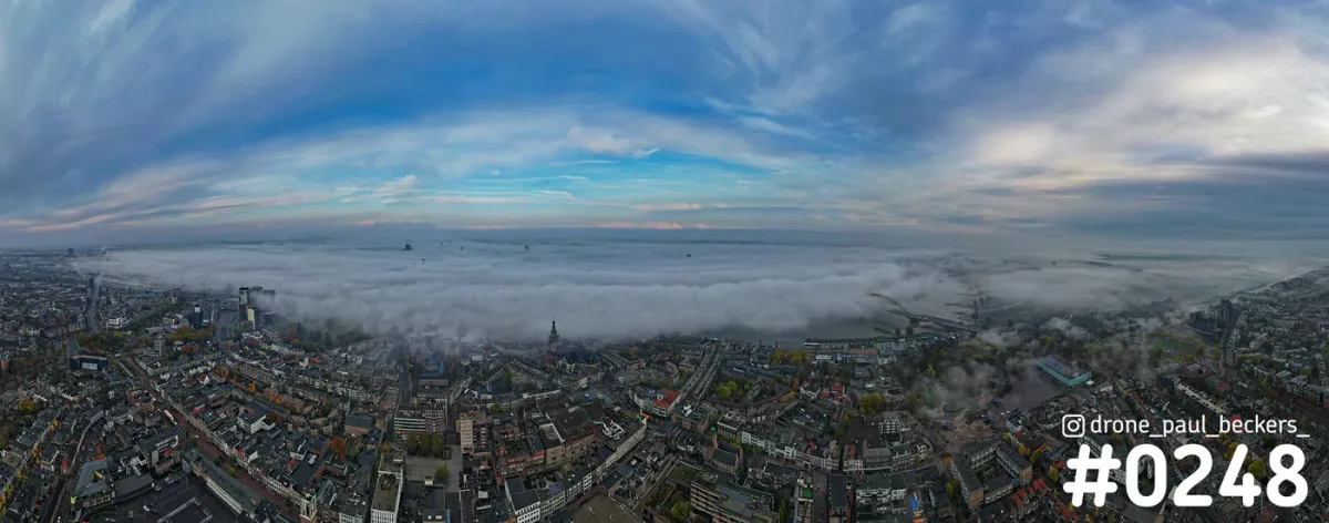Panoramafoto van Nijmegen met de Stevenskerk en een dik mistdek boven de Waal, onder een blauwe lucht met wolken