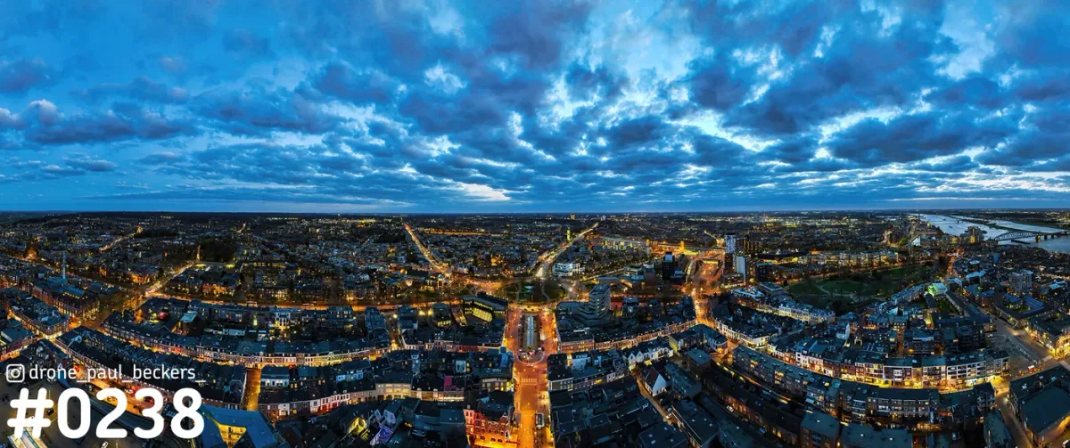 Panorama-luchtfoto van Nijmegen in het blauwe uur, met verlichte straten en de Waal rechts onder een wolkendek