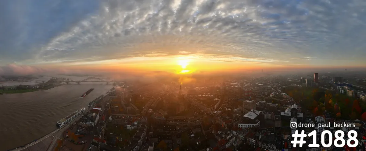 Panorama-luchtfoto van Nijmegen bij zonsopkomst, met de Waal links, de Waalbrug in de mist en een wolkenlucht erboven