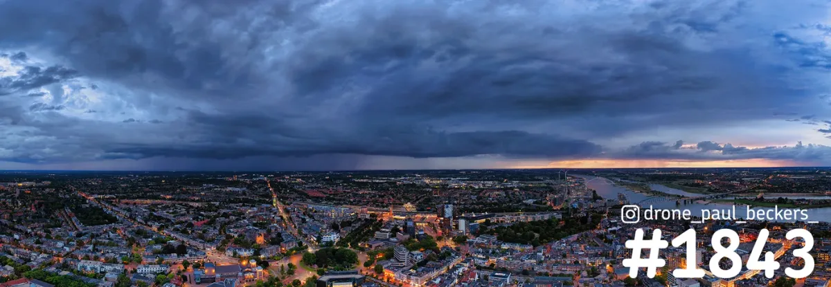 Panorama-luchtfoto van Nijmegen in de avondschemering, met zware donkere wolken boven de stad en de Waal rechts