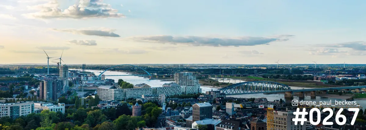 Luchtpanorama van Nijmegen met de rivier de Waal, een stalen boogbrug en windturbines in zacht avondlicht