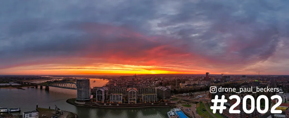 Panorama-luchtfoto van Nijmegen met de Nimbus aan de Waal en een rood-oranje gloed aan de horizon onder dikke wolken