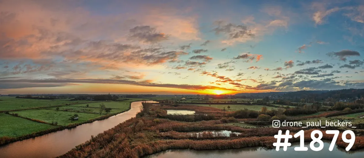 Luchtpanorama van de Ooijpolder bij zonsondergang, met een kronkelende waterloop, rietkragen en een kleurrijke wolkenlucht