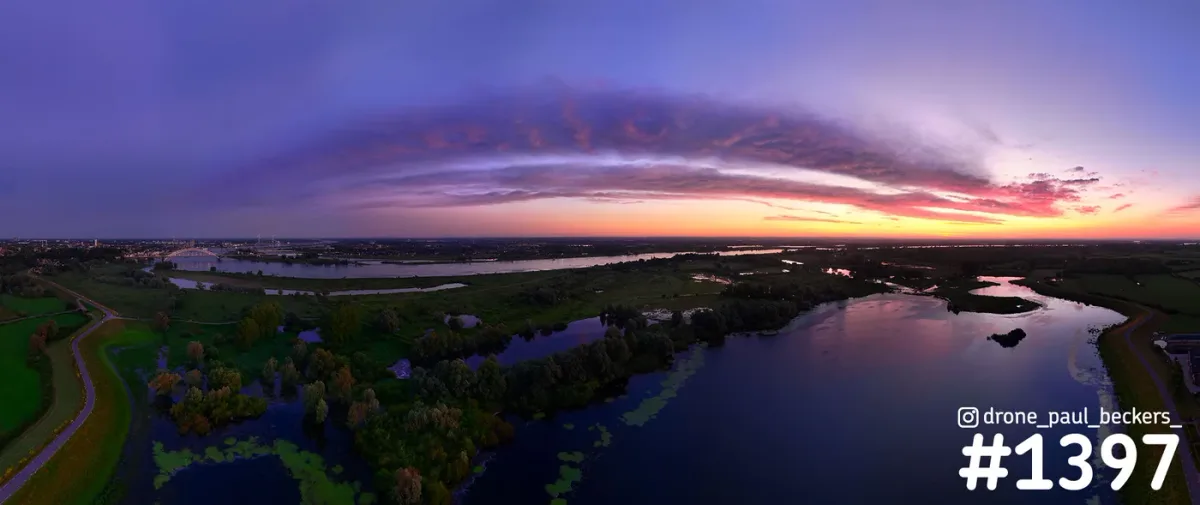 Panoramische luchtfoto van de Ooijpolder bij Nijmegen in de schemering, met paarsroze wolkenboog boven de Waal en spiegelende plassen