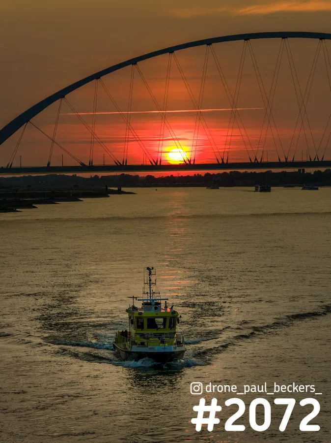 Luchtfoto van De Oversteek in Nijmegen bij zonsondergang, met een geelgroene boot op de Waal en de zon onder de boog