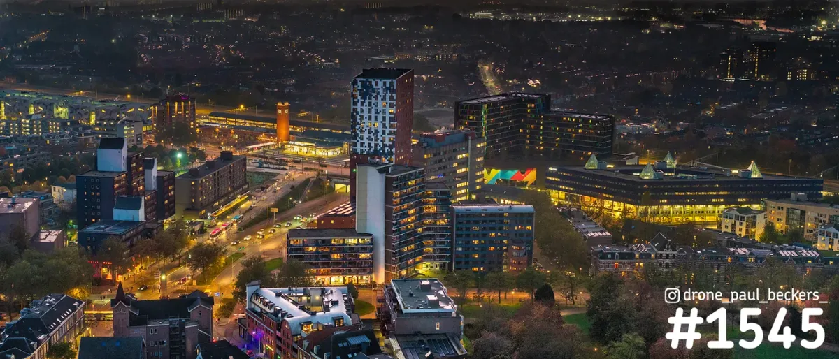 Luchtfoto van het Stationsgebied in Nijmegen in de avondschemering, met verlichte hoogbouw en kantoren rond het centrum
