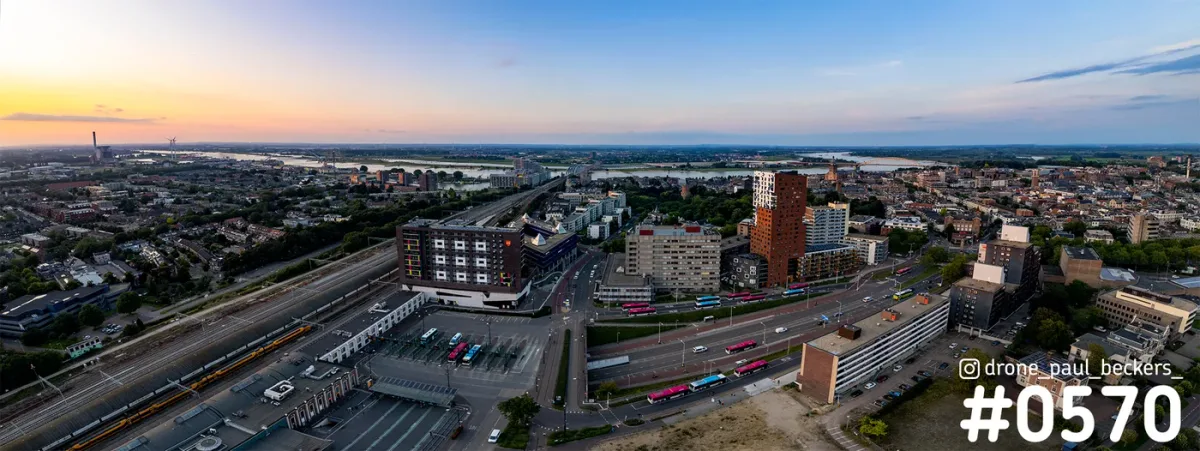 Panorama-luchtfoto van het Stationsgebied in Nijmegen in de schemering, met bussen, spoor en een rivier aan de horizon