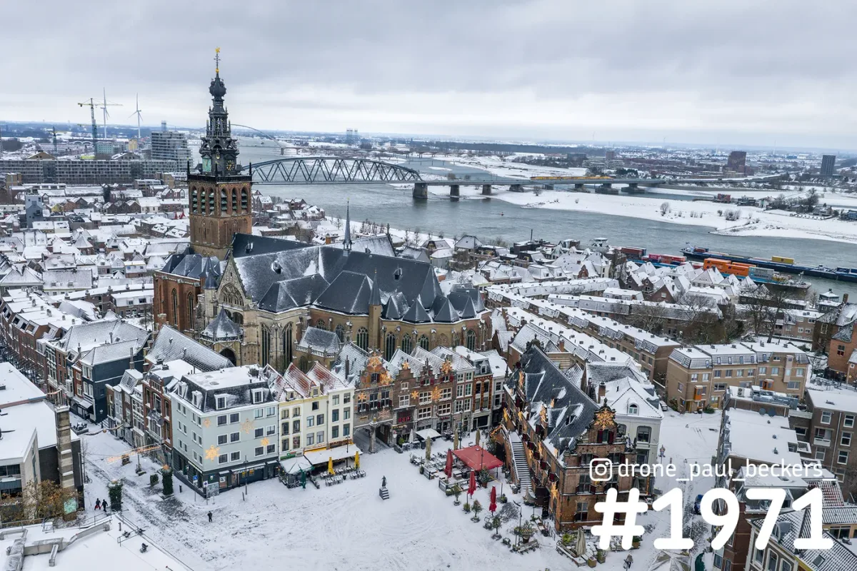 Luchtfoto van de Stevenskerk in Nijmegen onder een laag sneeuw, met de Grote Markt en de brug over de Waal