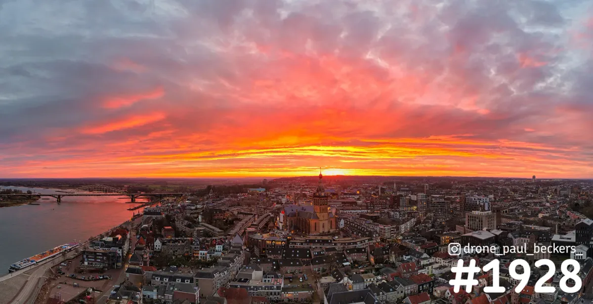 Luchtpanorama van Nijmegen met de Stevenskerk, de Waal en Waalbrug onder een vuurrode lucht vol wolken