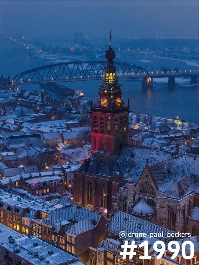 Luchtfoto van de Stevenskerk in Nijmegen met rood verlichte toren en besneeuwde daken, met de Waal en een brug erachter