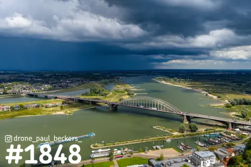 Luchtfoto van de Waalbrug in Nijmegen met donkere regenwolken boven de Waal en de uiterwaarden bij Lent