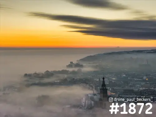 Luchtfoto van de Stevenskerktoren in Nijmegen die boven de mist uitsteekt, met oranje zonsopkomst en donkere wolkenbanden