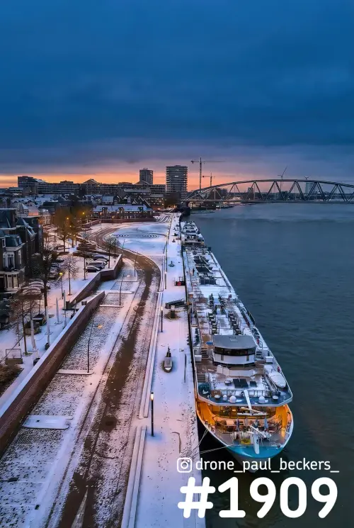 Luchtfoto van de Waalkade in Nijmegen met sneeuw, een afgemeerd rivierschip op de Waal en een oranje gloed bij avondschemer