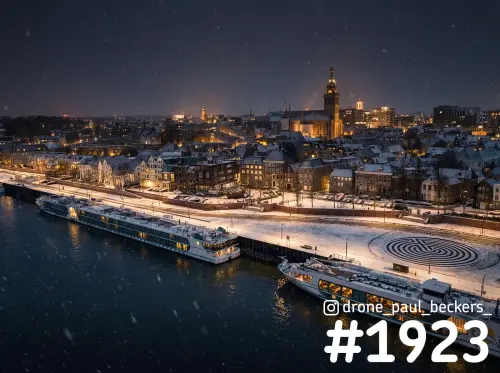 Luchtfoto van de Waalkade in Nijmegen in de sneeuw, met twee rivierschepen aan de kade en de verlichte Stevenskerk erachter
