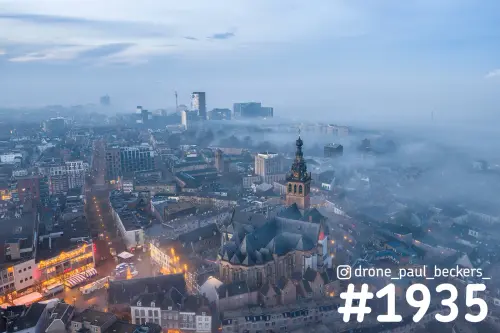 Luchtfoto van de Stevenskerk in Nijmegen in blauwe schemering, met mist tussen de daken en hoogbouw op de achtergrond