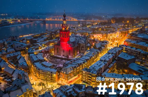 Luchtfoto van de rood verlichte Stevenskerk in Nijmegen in de sneeuw, met de Waalbrug en de Waal op de achtergrond