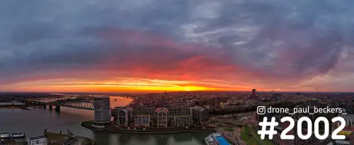 Panorama-luchtfoto van Nijmegen met de Nimbus aan de Waal en een rood-oranje gloed aan de horizon onder dikke wolken