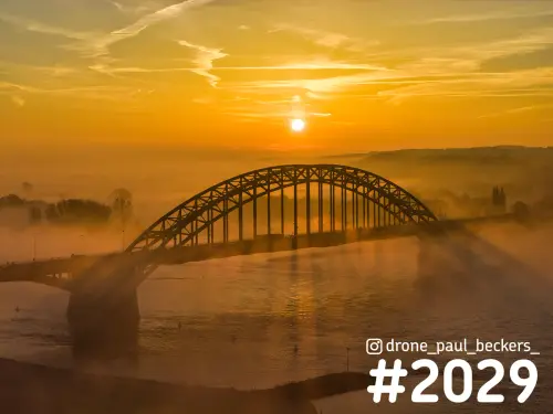 Luchtfoto van de Waalbrug in Nijmegen bij zonsopkomst, met de zon boven de boogbrug en mist die over de Waal trekt