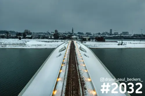Luchtfoto van een besneeuwde brug over de nevengeul bij Lent, met de Stevenskerk van Nijmegen in de grijze winterlucht