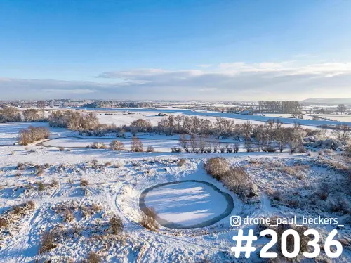 Luchtfoto van de besneeuwde Ooijpolder bij Nijmegen met een bevroren poel in de voorgrond onder een blauwe winterlucht