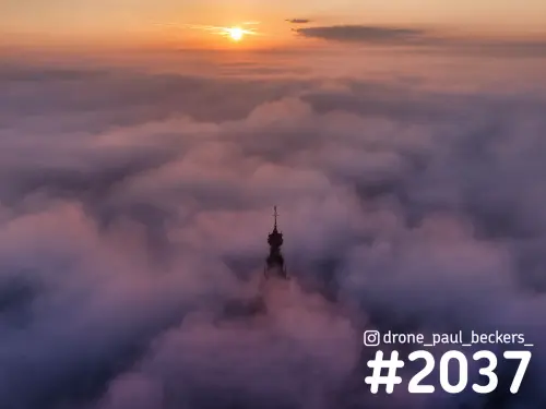Luchtfoto van de torenspits van de Stevenskerk in Nijmegen die boven een dik mistdek uitsteekt, met zonsopkomst aan de horizon