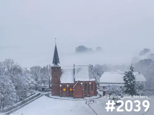 Luchtfoto van een rode baksteenkerk in de Ooijpolder in de sneeuw, met verlichte ramen en mist boven het landschap