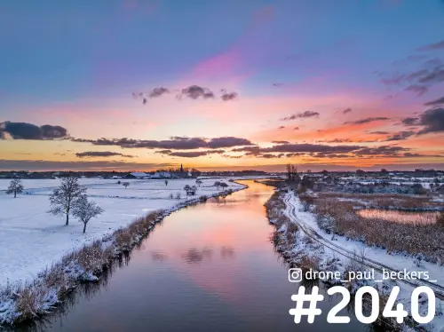Luchtfoto van een besneeuwde Ooijpolder in de winter, waterloop met roze schemerlicht en kale bomen aan de oever