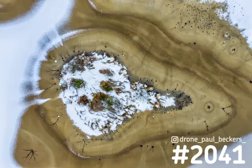 Luchtfoto van een bevroren ven in de Hatertse Vennen met een besneeuwd eilandje en gouden ijsringen eromheen