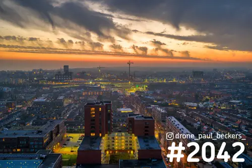 Luchtfoto van het Stationsgebied in Nijmegen bij schemering, met verlichte flatgebouwen en een oranje wolkenlucht