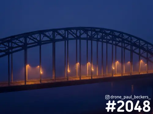 Waalbrug in Nijmegen in de blauwe avondschemering, met oranje straatlantaarns die door de mist boven de Waal gloeien