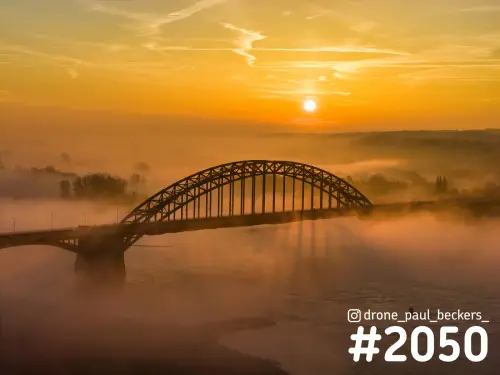 Luchtfoto van de Waalbrug in Nijmegen bij zonsopkomst, met laaghangende mist boven de rivier de Waal en een oranje lucht