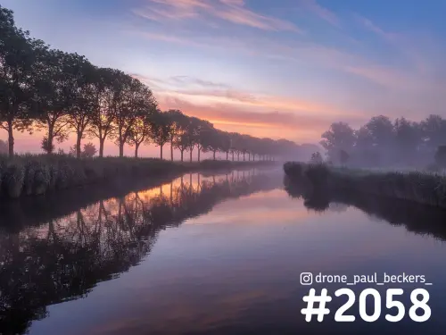 Luchtfoto van een rij bomen langs een rustig water in de Ooijpolder bij zonsopkomst, met mist en roze lucht weerspiegeld