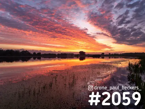 Luchtfoto van de Ooijpolder bij zonsopkomst, met oranjerode wolken die weerspiegelen in ondiep water met riet en waterplanten