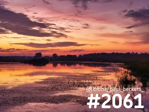 Luchtfoto van de Ooijpolder bij zonsopkomst, met oranje en paarse wolken die weerspiegelen in een ondiepe plas met waterplanten