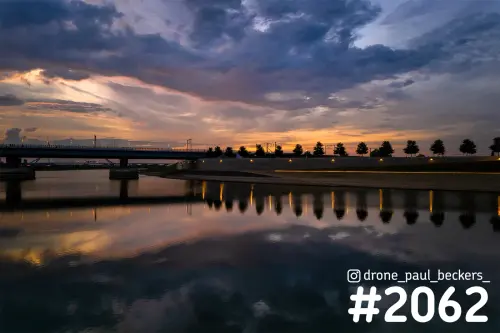 Luchtfoto van de nevengeul bij Lent in Nijmegen met brug, spiegelend water en oranje gloed onder donkere wolken