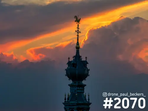 Torenspits van de Stevenskerk in Nijmegen met haan als windvaan, afgetekend tegen oranje wolken