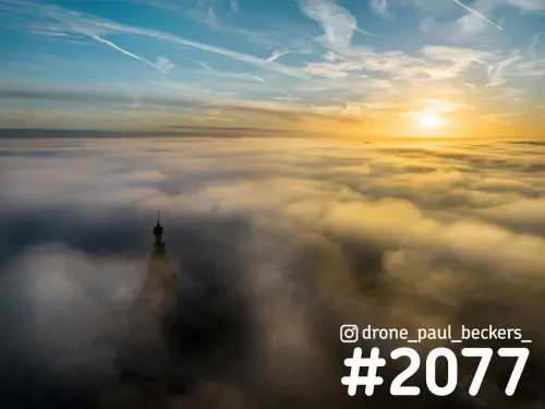 Luchtfoto van de Stevenskerk in Nijmegen die als silhouet uit een dik mistdek steekt, met laagstaande zon aan de horizon