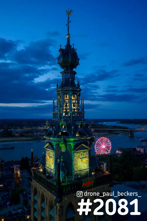 Luchtfoto van de Stevenskerk in Nijmegen in het blauwe uur, met groen verlichte toren en een reuzenrad bij de Waal